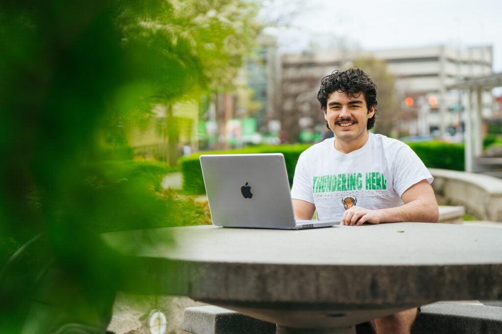 student sitting at a table outside the library with a laptop and smiling at the camera