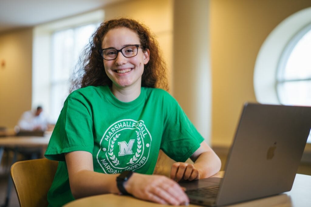 student working on a laptop in the library and smiling at the camera