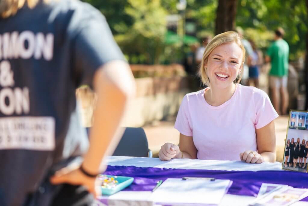 student smiling at the camera sitting at a table outside on campus, talking with another student