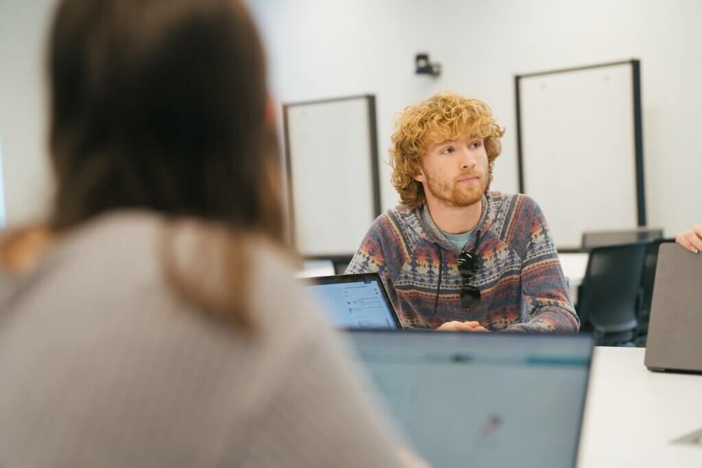 two students sitting across from each other in a classroom on laptops