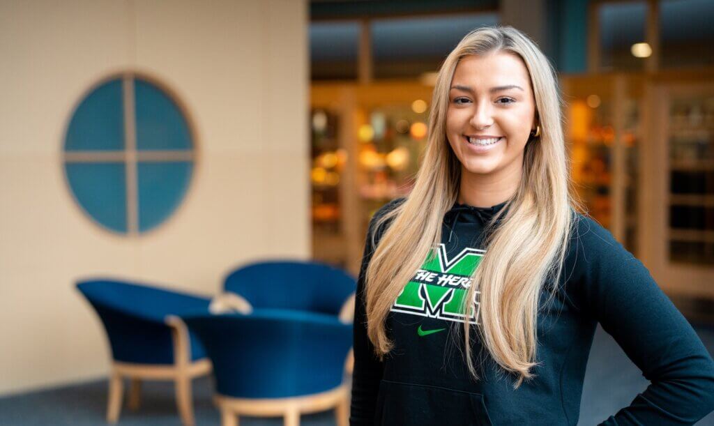 student in the library on campus, smiling at the camera