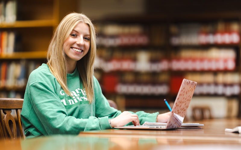 A student smiles at the camera while working on a laptop in a library