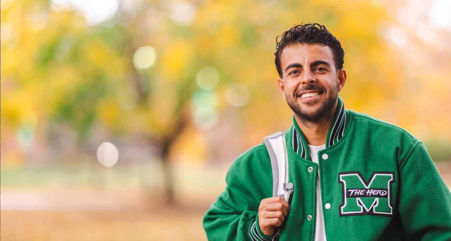 A student wearing a Marshall University letter jacket, holding the strap of a backpack, stands in front of a yellow tree on Marshall University's Huntington campus