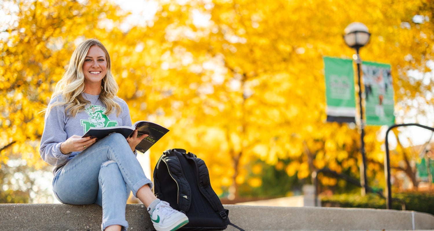 A student reads on the plaza in front of fall trees on Marshall University's Huntington campus