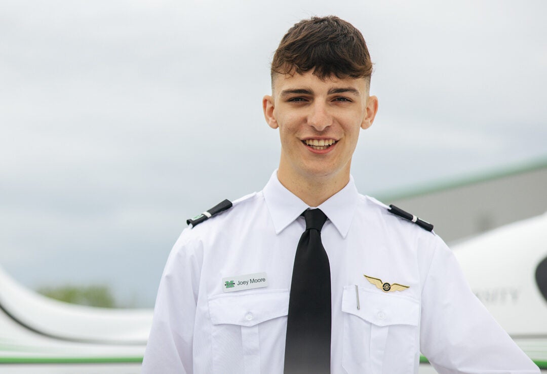 A Marshall University Bill Noe Flight School student wears a pilot uniform while standing in front of a Marshall branded aircraft.