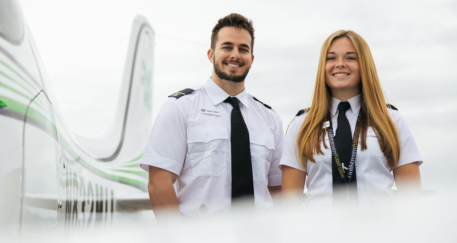 Two students, wearing pilot uniforms, from Marshall University's Bill Noe Flight School smile in front of a Marshall branded airplane.