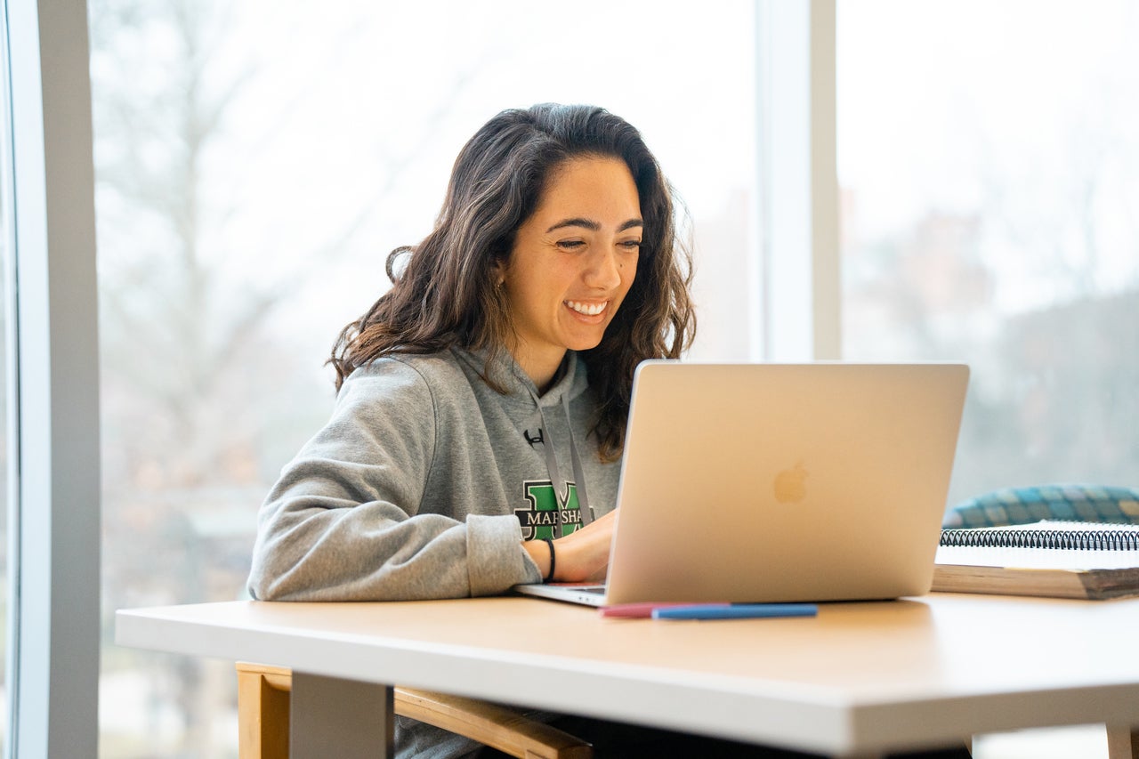Marshall student sits near a window on a laptop