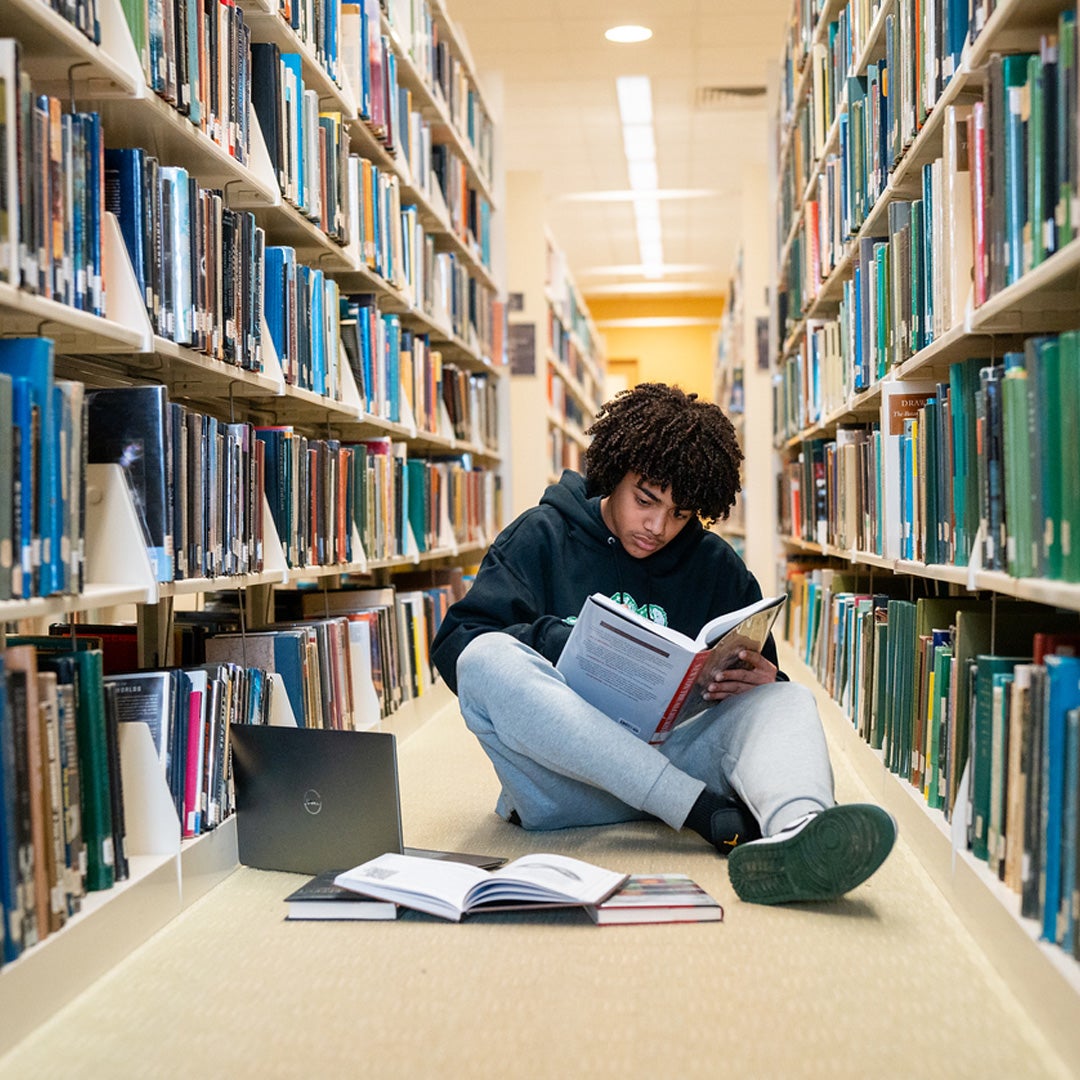 A student sits on the floor of the library with a laptop, surrounded by books.