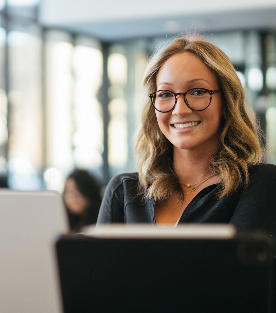 A student smiles at the camera while on a laptop in a library