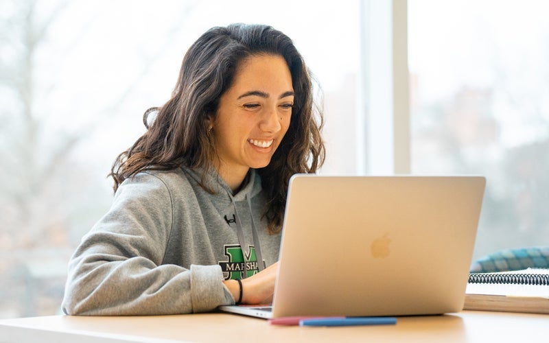 A student in a Marshall sweatshirt smiles at a computer screen