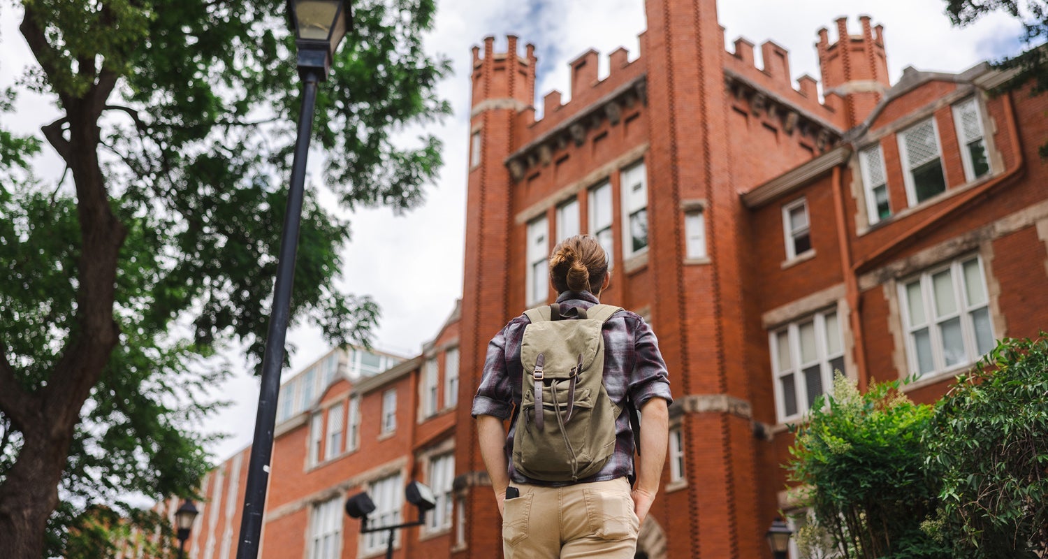 A student wearing a backpack looks up at Marshall University's Old Main from the sidewalk