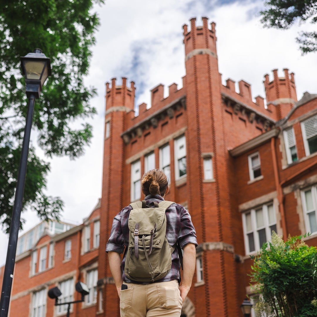 A student wearing a backpack looks up at Marshall University's Old Main from the sidewalk