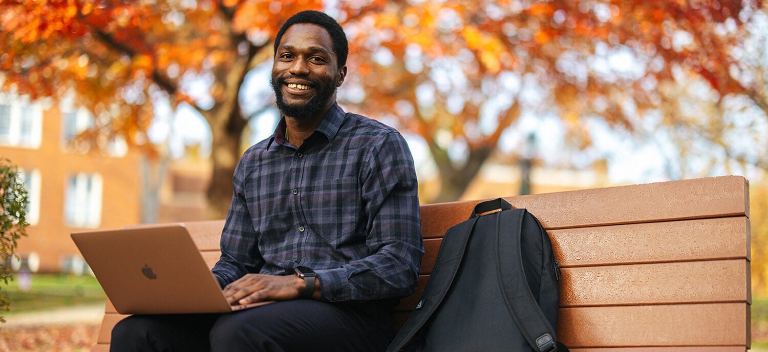 Student with a laptop, on a bench surrounded by fall colors