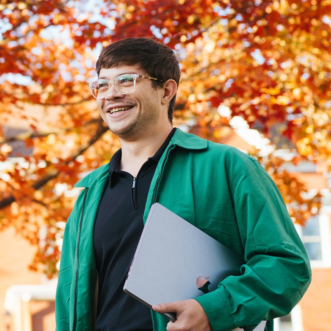 Student walking in front of a tree full of orange leaves while carrying a laptop