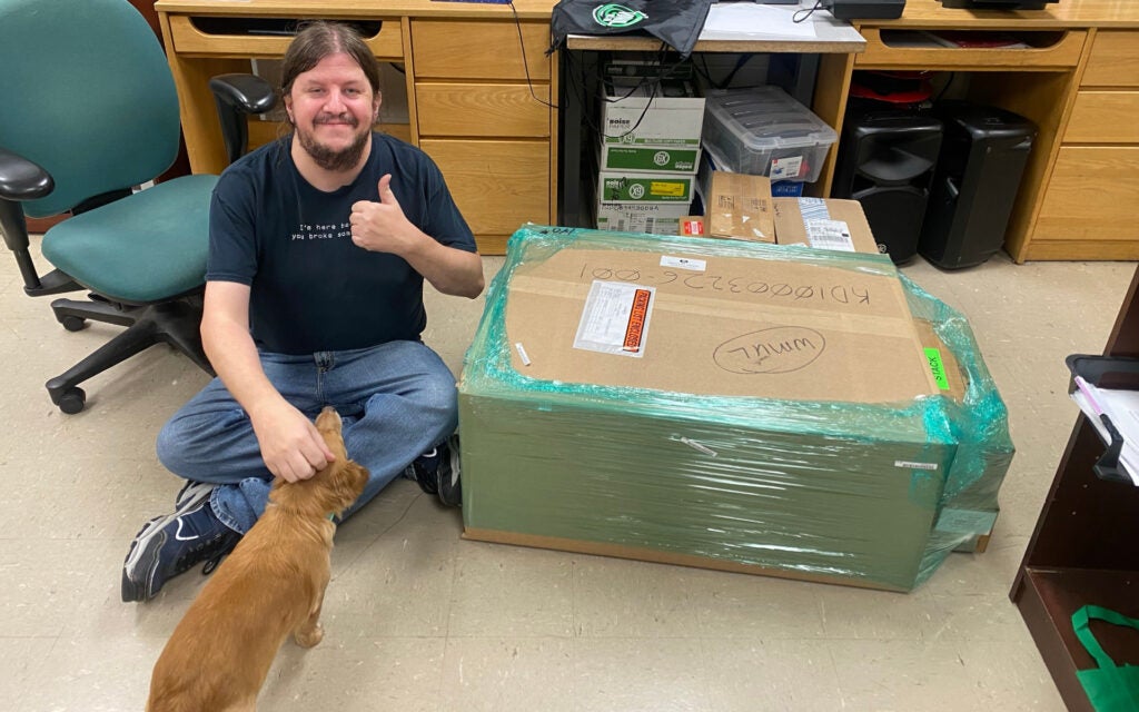 Mike Stanley sits next to the newly delivered transmitter. Gaia Wheeler, station mascot, is helping.