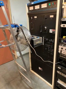 The exciter has been slid out from inside the transmitter and the top cover has been removed. A floor fan is attached to a step ladder and is blowing onto the exciter.